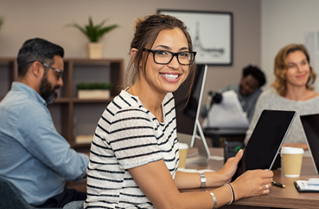 woman smiling while using tablet. Man and women in background also using computer for classes online.